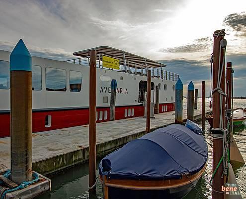 The white and red hotel barge "Ave Maria" for Bike & Barge tours moored at a pier in the Venice Lagoon, with a covered boat in the foreground.