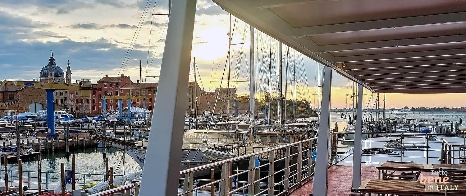 View from a ship's deck over Venice harbor at sunrise, featuring a classic motorboat and the city skyline in the background.