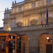 Milanese Charm: Teatro alla Scala and the Historic Tram • m24o The illuminated facade of Teatro alla Scala in Milan at dusk, with a historic yellow tram passing by in the foreground.