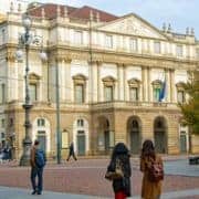 Teatro alla Scala Milan – Legendary Opera House • m24o Two young women walking across Piazza della Scala in Milan with the Teatro alla Scala in the background under a clear sky.