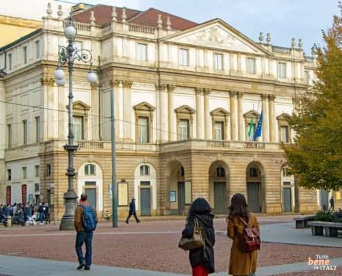 Teatro alla Scala Milan – Legendary Opera House • m24o Two young women walking across Piazza della Scala in Milan with the Teatro alla Scala in the background under a clear sky.