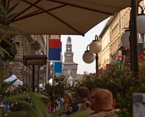 Via Dante Milan – View towards Castello Sforzesco • m24o Perspective view of the pedestrian street Via Dante in Milan, featuring the Filarete Tower of Castello Sforzesco framed by historic buildings in the background.