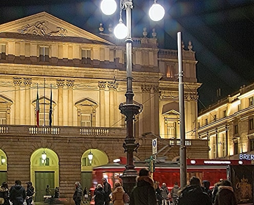 The illuminated Teatro alla Scala in Milan at night with pedestrians in the piazza and a passing red tram in the foreground.