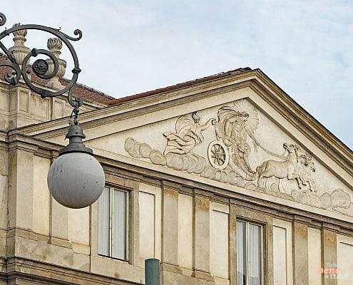 Architectural Art: Apollo’s Chariot at La Scala • m24o Close-up of the pediment of Teatro alla Scala in Milan featuring the "Chariot of Apollo" relief and a historic street lamp in the foreground.