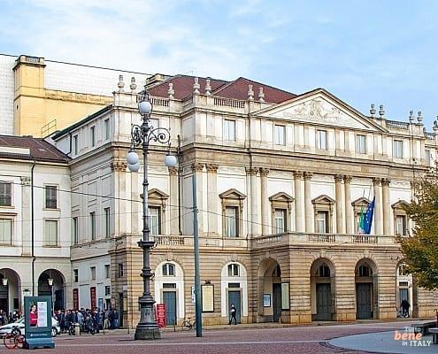 A Front-Row Seat in the City: Teatro alla Scala from the Piazza • m24o Panoramic exterior view of Teatro alla Scala in Milan featuring its neoclassical facade and the modern stage tower under a blue sky.