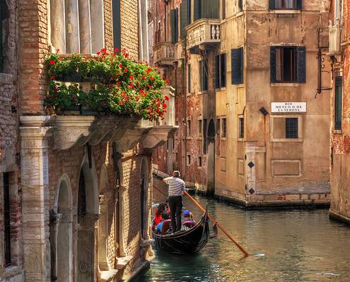 Pure Romance: A Gondola Ride Through Venice's Hidden Canals • ©Photocreo Bednarek - adobe A gondolier steers his gondola with passengers through a narrow, romantic canal in Venice, passing historic buildings with flower-filled balconies; Photo © Photocreo Bednarek - Adobe Stock.