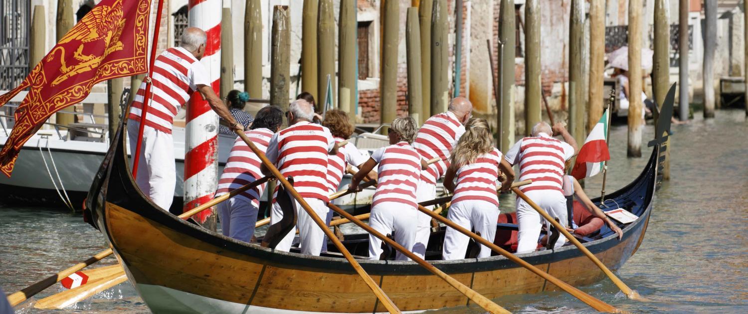 A crew of eight rowers in red and white striped jerseys steering a traditional Venetian "Bissona" boat through a canal; Photo &copy; Mauro Dalla Pozza - Adobe Stock.