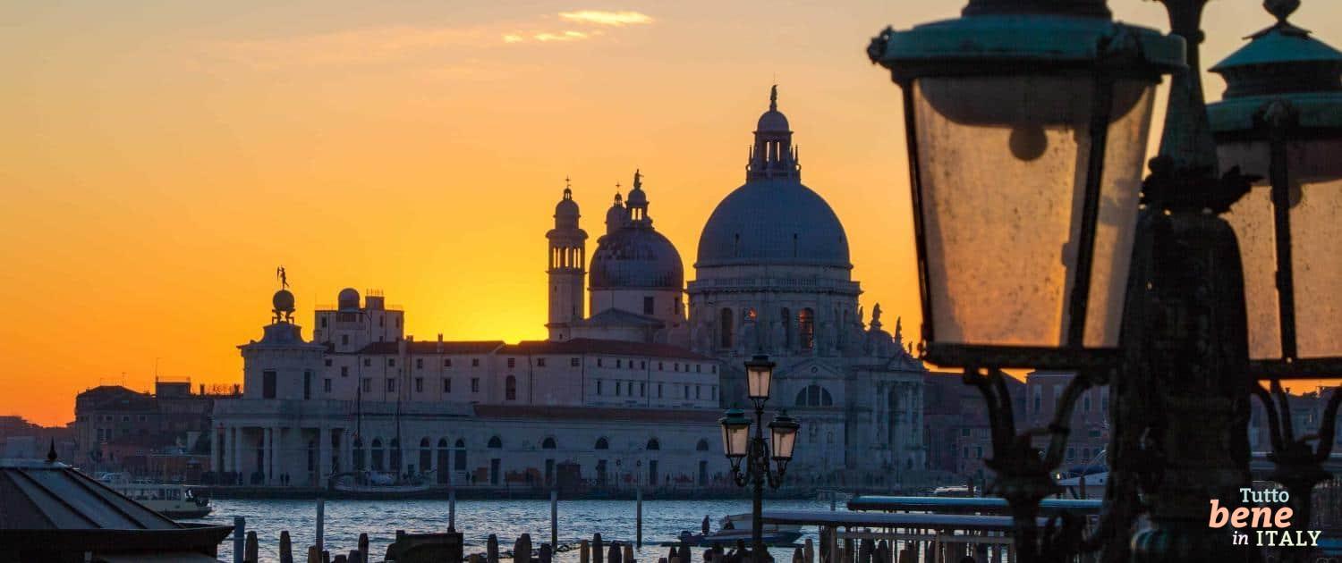 Silhouette der Basilika Santa Maria della Salute in Venedig bei einem leuchtend orangefarbenen Sonnenuntergang, eingerahmt von historischen Straßenlaternen im Vordergrund.
