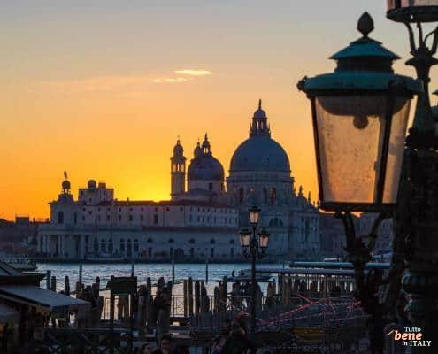 Abendstimmung in Venedig – Sonnenuntergang über der Salute-Kirche • m24o Silhouette der Basilika Santa Maria della Salute in Venedig bei einem leuchtend orangefarbenen Sonnenuntergang, eingerahmt von historischen Straßenlaternen im Vordergrund.