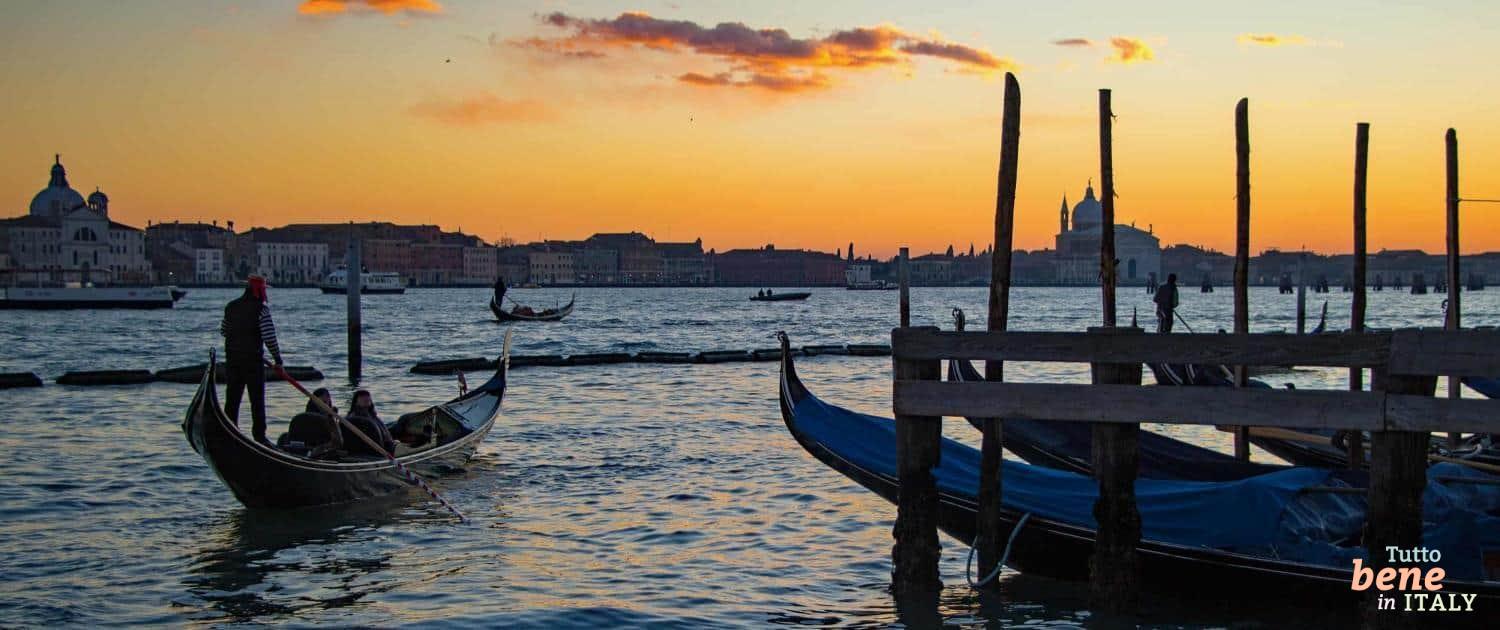 Ein Gondoliere steuert seine Gondel bei einem leuchtend orangefarbenen Sonnenuntergang auf dem Canal Grande in Venedig; im Hintergrund die Silhouette der Stadt.