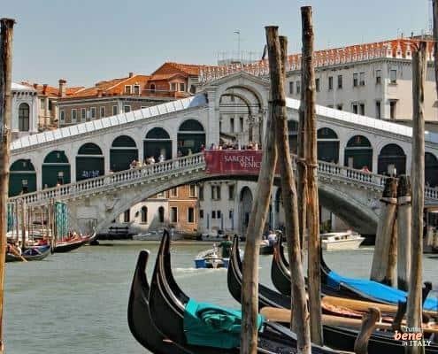 View through wooden gondola poles of the historic Rialto Bridge on the Grand Canal in Venice under a clear blue sky.