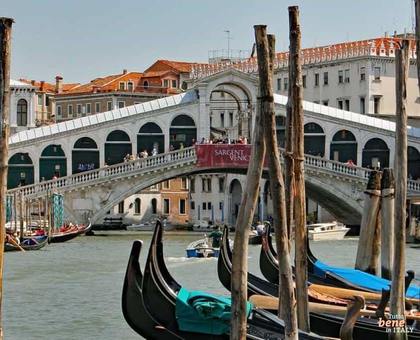 Das Herz von Venedig: Die Rialtobrücke über den Canal Grande • m24o Blick durch die hölzernen Gondelstangen auf die historische Rialtobrücke über den Canal Grande in Venedig unter strahlend blauem Himmel.