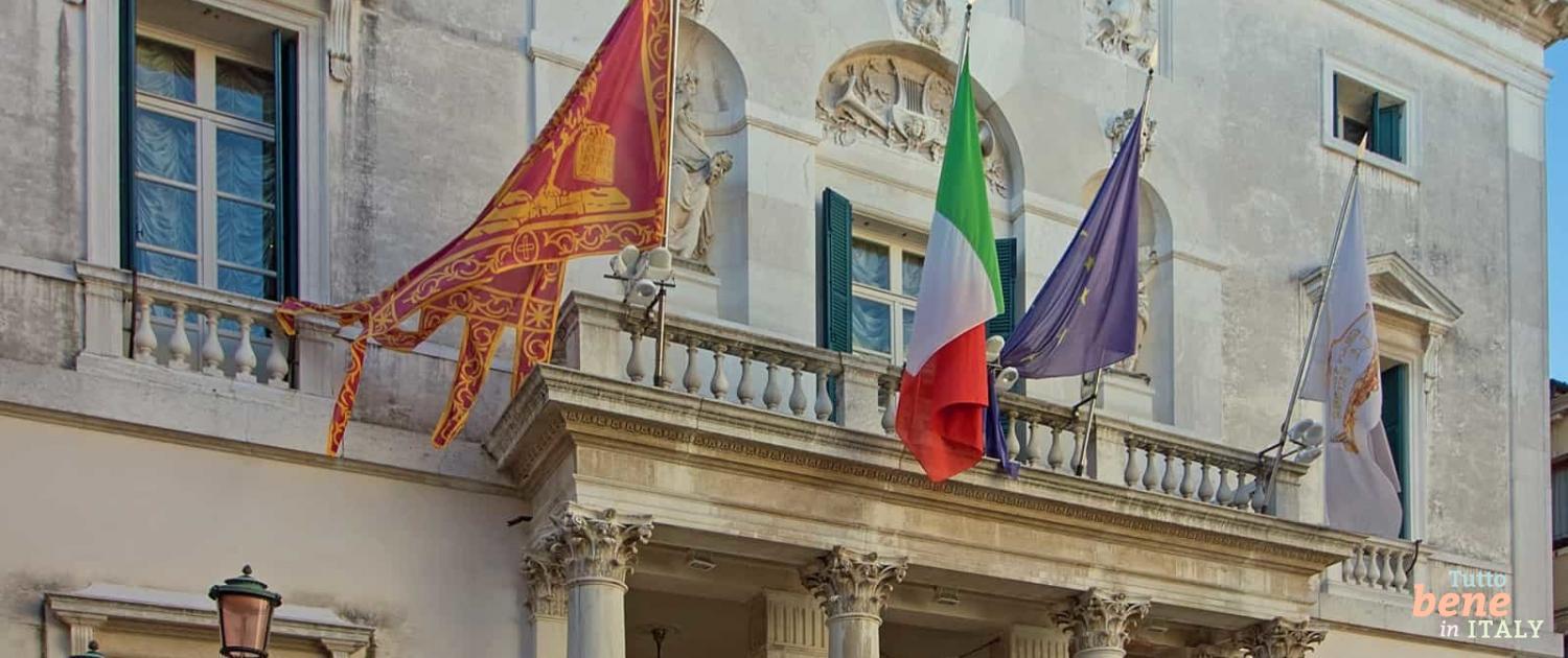 The magnificent facade of Teatro La Fenice in Venice featuring fluttering flags and neoclassical sculptures under a clear blue sky.