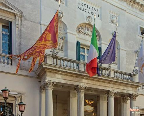 The magnificent facade of Teatro La Fenice in Venice featuring fluttering flags and neoclassical sculptures under a clear blue sky.