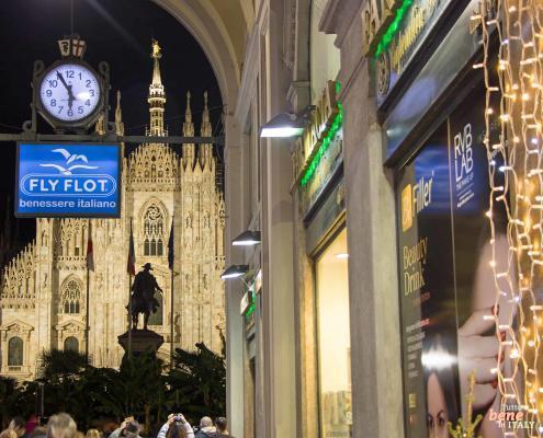 A nighttime view of the illuminated Milan Cathedral, framed by a stone archway with a hanging clock and lit shop windows in the foreground.