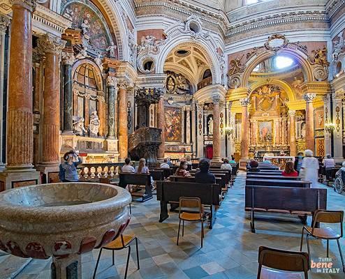 Interior of the Royal Church of San Lorenzo in Turin • m24o Magnificent baroque interior of the San Lorenzo Church in Turin with churchgoers seated in wooden pews, a large marble holy water font in the foreground, highly ornate marble columns, and gilded altars under complex arched architecture.