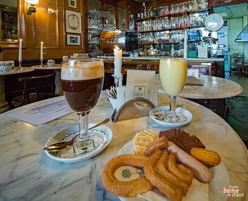 Traditional Bicerin drink at the historic Caffè Al Bicerin in Turin • m24o A glass of traditional Turin Bicerin drink showing layered espresso, chocolate, and cream, alongside a plate of Piedmontese pastries on a marble table inside a historic wooden café.