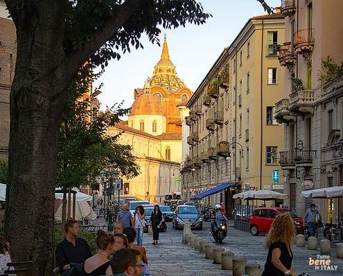 Blick auf die Kapelle des Turiner Grabtuchs von der Via Palazzo di Città • m24o Blick durch eine belebte Turiner Straße mit Café-Gästen im Schatten eines Baumes links, einer Radfahrerin rechts und der markanten, von der Abendsonne beleuchteten Kuppel der Grabtuchkapelle im Hintergrund.