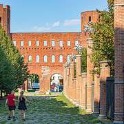 Porta Palatina Turin Roemisches Stadttor Antike Ruine Backstein • m24o Ein Paar spaziert auf einem von Bäumen und Ziegelpfeilern gesäumten Weg auf das antike römische Stadttor Porta Palatina in Turin zu.