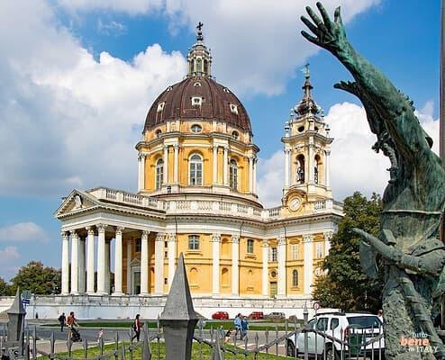 Basilica of Superga in Turin with Monument • m24o The baroque Basilica of Superga in Turin featuring its large brown dome and yellow-and-white facade under a partly cloudy sky, with a bronze statue with raised arms in the right foreground.