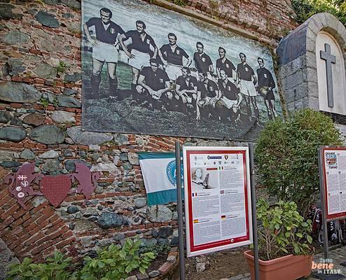 Gedenkstätte des Grande Torino in Superga mit Infotafeln • m24o Die Gedenkstätte für die Fußballmannschaft Grande Torino an einer rustikalen Steinmauer in Superga mit zwei Infotafeln auf Ständern im Vordergrund, einem großen historischen Mannschaftsfoto und einer steinernen Grabstätte mit Kreuz im Hintergrund.