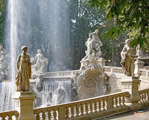 The monumental Fontana dei 12 Mesi fountain in Turin's Valentino Park, featuring a tall geyser water jet, cascading waterfalls, multiple classical stone statues on pedestals, and an ornate balustrade staircase in the foreground.