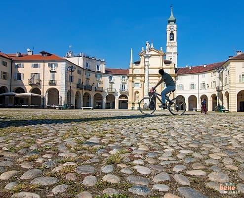 Piazza dell'Annunziata in Venaria Reale bei Turin mit Radfahrer • m24o Ein Fahrradfahrer überquert die von historischen, geschwungenen Gebäuden gesäumte Piazza dell'Annunziata in Venaria Reale auf grobem Kopfsteinpflaster, im Hintergrund die barocke Kirche Chiesa della Natività di Maria Vergine unter blauem Himmel.