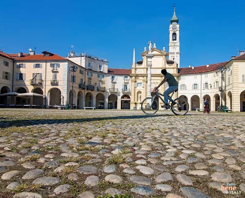 Piazza dell'Annunziata in Venaria Reale bei Turin mit Radfahrer • m24o Ein Fahrradfahrer überquert die von historischen, geschwungenen Gebäuden gesäumte Piazza dell'Annunziata in Venaria Reale auf grobem Kopfsteinpflaster, im Hintergrund die barocke Kirche Chiesa della Natività di Maria Vergine unter blauem Himmel.