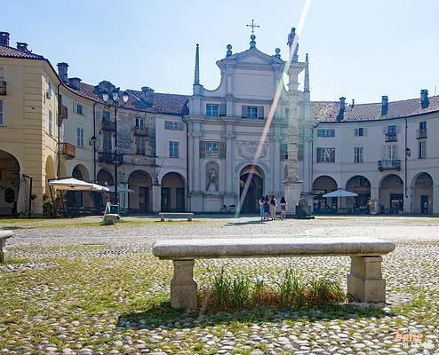 Piazza dell'Annunziata in Venaria Reale with Stone Bench in Foreground • m24o A rustic stone bench in the foreground on the historic cobblestone pavement of Piazza dell'Annunziata in Venaria Reale, with the baroque church facade, a tall stone column, pedestrians, and curved palace buildings in the background under a blue sky with sun flares.