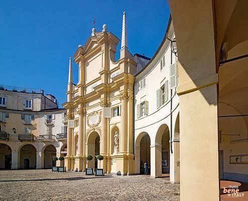 Piazza dell'Annunziata und Chiesa della Natività di Maria Vergine in Venaria Reale • m24o Blick aus den schattigen Arkaden auf die geschwungene Piazza dell'Annunziata in Venaria Reale mit der hellgelben barocken Kirchenfassade, verzierten Säulen und kleinen Bäumen in Kübeln vor einem gepflasterten Platz unter blauem Himmel.