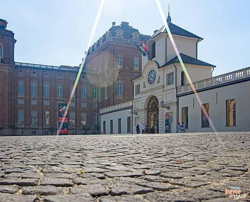 Ehrenhof von Schloss Venaria Reale bei Turin, Weltkulturerbe der Unesco • m24o Blick aus Bodennähe über das historische Kopfsteinpflaster des Ehrenhofs von Schloss Venaria Reale in Turin mit dem hellen Uhrenturm rechts, den monumentalen Backsteinfassaden im Hintergrund und Sonnenstrahlen, die ins Bild fallen.