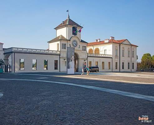 Eingangsbereich von Schloss Venaria Reale bei Turin • m24o Der historische Uhrenturm und die barocken Palastflügel des Schlosses Venaria Reale an einem weiten, gepflasterten Platz unter klarem blauen Himmel mit vereinzelten Besuchern im Hintergrund.