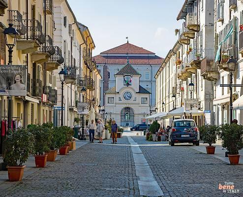 Fußgängerzone Via Andrea Mensa in Venaria Reale bei Turin • m24o Blick durch eine gepflasterte Einkaufsstraße in Venaria Reale mit historischen Wohnhäusern, Balkonen, Topfpflanzen und Passanten, die auf den barocken Uhrenturm des Schlosses am Ende der Straße zulaufen.