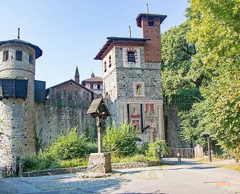Entrance Gate to the Borgo Medievale in Valentino Park in Turin • m24o The stone fortress walls of the Borgo Medievale in Turin featuring a round watchtower on the left, a tall gate tower with frescoes on the right, a wooden wayside cross in the foreground, and pedestrians under a blue sky.