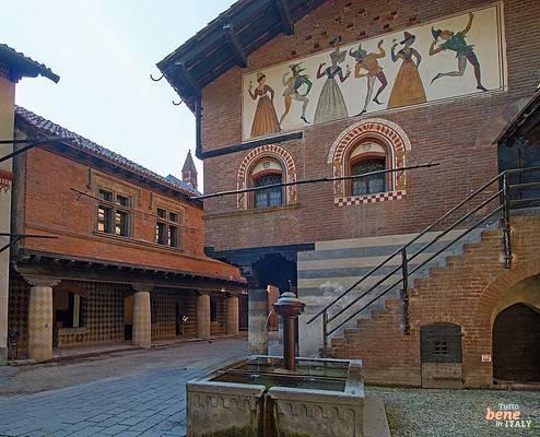 Courtyard at the Borgo Medievale in Valentino Park in Turin • m24o A historic courtyard inside the Borgo Medievale in Turin with a stone fountain in the foreground, a brick building facade decorated with frescoes of dancing figures on the upper right, and porticoes in the background.