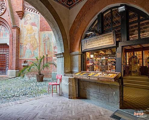 Handwerkswerkstatt und Kirche im Borgo Medievale in Turin • m24o Blick unter einem gemauerten Bogen auf eine historische Handwerkswerkstatt mit Schaukasten rechts und die gotische Backsteinfassade einer Kirche mit bunten Heiligenfresken und einer Topfpalme links auf einem Pflasterhof.