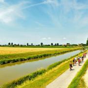 A group of cyclists riding on a paved path alongside a canal in the flat landscape of Northern Italy under a bright blue sky; Photo © Girolibero.