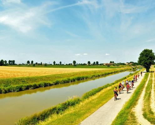 A group of cyclists riding on a paved path alongside a canal in the flat landscape of Northern Italy under a bright blue sky; Photo © Girolibero.