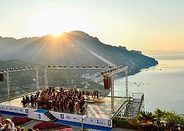 A symphony orchestra performs at the Ravello Festival on the Villa Rufolo Belvedere stage as the sun rises brightly over the Amalfi Coast mountains.
