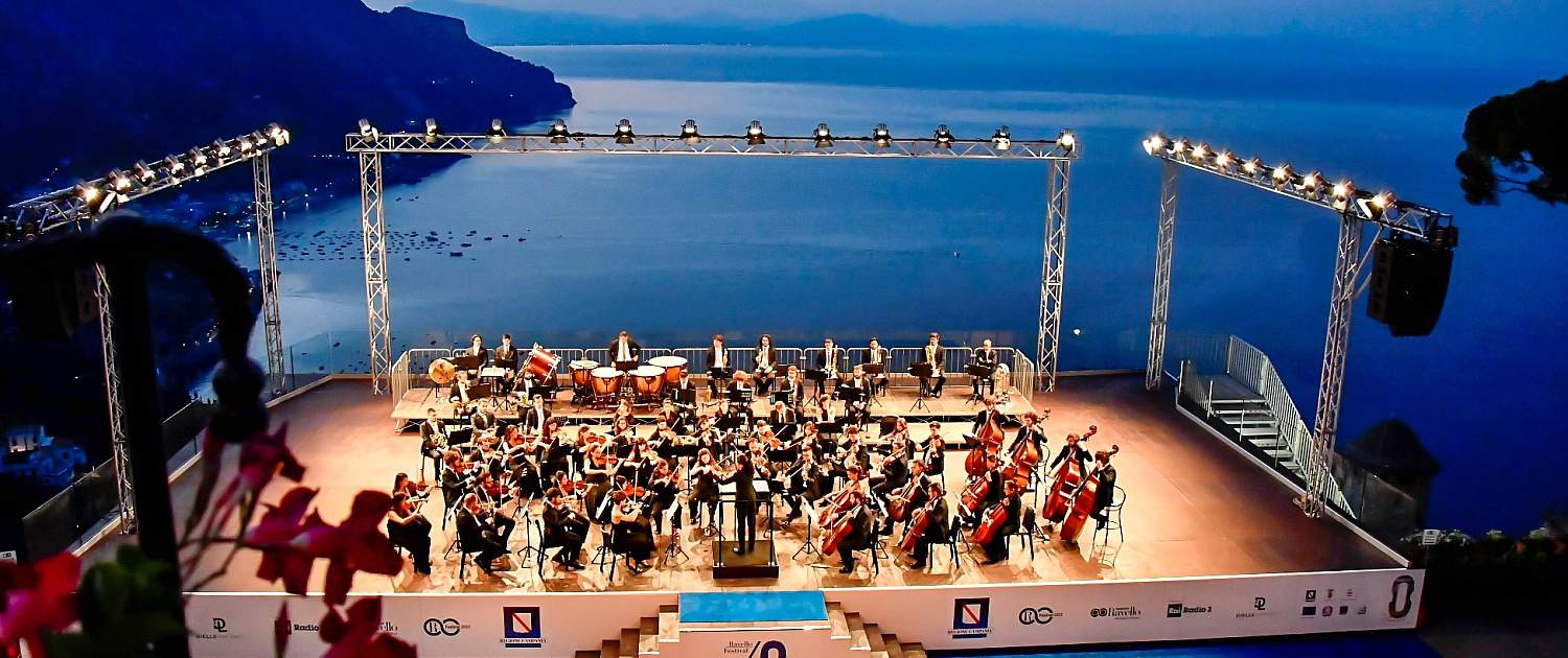 Symphony orchestra on the Villa Rufolo Belvedere stage during the Ravello Festival at dusk overlooking the sea.