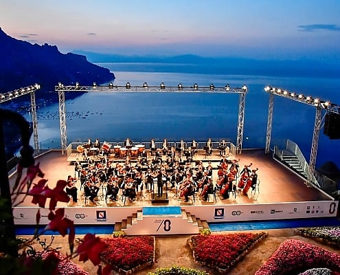 Symphony orchestra on the Villa Rufolo Belvedere stage during the Ravello Festival at dusk overlooking the sea.