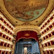 The opulent auditorium of Teatro di San Carlo featuring six tiers of gold-decorated boxes, a large ceiling fresco, and the stage with a red velvet curtain.