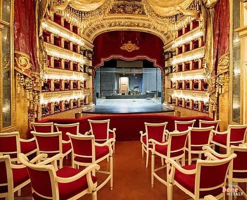 The opulent golden auditorium of Teatro di San Carlo with red tiers and white chairs in the foreground, viewed from the elevated Royal Box looking towards the stage.