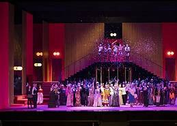 Cast and choir in elegant costumes on a vibrant red lit opera stage at Teatro Regio di Torino with falling confetti.
