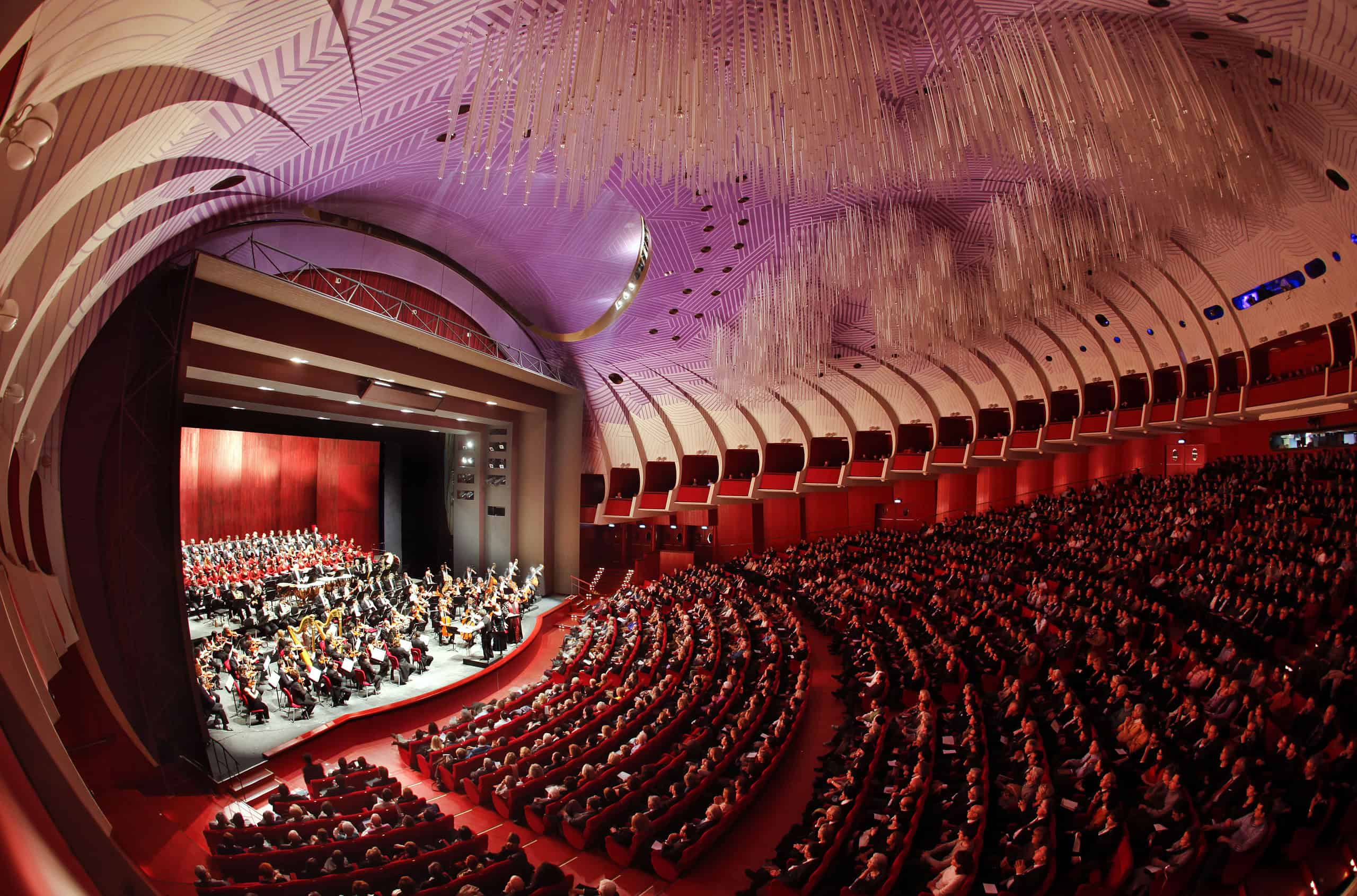 Wide view of the stage with orchestra and choir inside the packed auditorium of Teatro Regio di Torino featuring its modern purple ceiling.