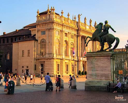 Palazzo Madama in Turin with Castor and Pollux Statues at Sunset • m24o The baroque Palazzo Madama and a medieval brick tower in Turin illuminated by golden evening light, framed by two large equestrian statues on stone pedestals in the foreground with pedestrians on the square.