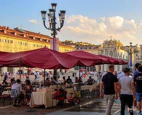 Piazza San Carlo in Turin at Sunset • m24o Bustling baroque square Piazza San Carlo in Turin with large red umbrellas of an outdoor café in the foreground, historic twin churches in the background, and pedestrians walking in warm evening light.