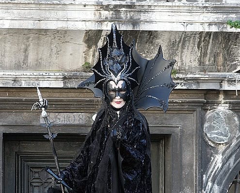 A mysterious figure at the Venice Carnival dressed in a magnificent black costume with a sparkling headpiece and scepter in front of a historic building.