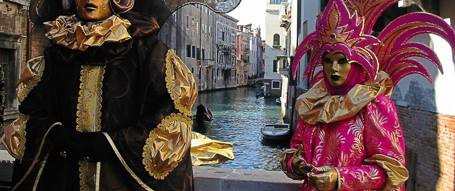 Zwei prächtig maskierte Gestalten in aufwendigen schwarzen und pinkfarbenen Barock-Kostümen auf einer Brücke über einem Kanal in Venedig.