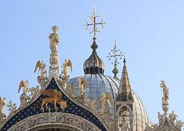 Upper facade of St. Mark’s Basilica in Venice with ornate statues, domes, golden decorations, and the winged lion symbol of St. Mark against a blue starry background.